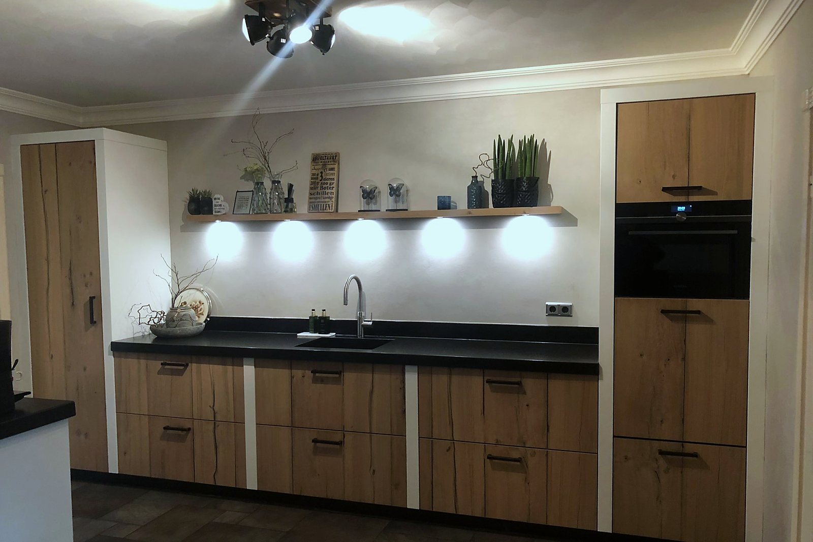 Eiken keuken langs de wand met zwart werkblad en verlichte nis boven het aanrecht, door Maatmakers Interieurbouw