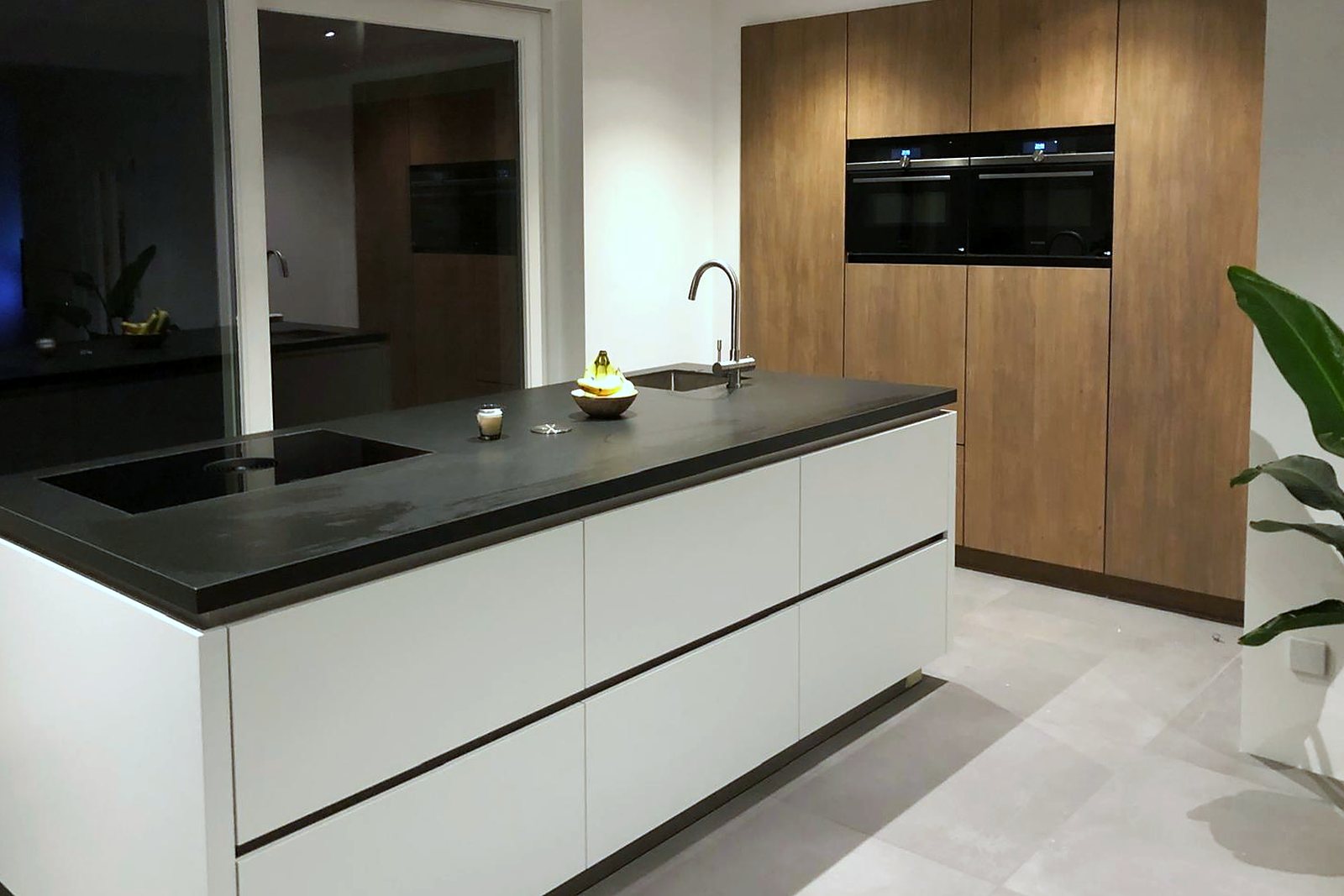 Greeploos wit kookeiland met donker werkblad tegenover een eiken kastenwand met inbouwapparatuur, door Maatmakers Interieurbouw